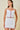 A woman modeling a white two-piece set consisting of a camisole top and a mini skirt, both featuring red scalloped trim and matching red piping details, set against a solid beige background.