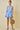 Woman wearing a light blue dress with floral patterns, holding a woven bag against a beige background