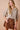 A woman modeling a brown and beige gingham long-sleeve blouse with a ruffled collar and button front, paired with a brown ruffled mini skirt, against a solid beige background.