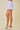 A person wearing lavender and white gingham mini shorts with a scalloped hem, paired with white heeled mule sandals, against a solid beige studio background.