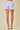 A person shown from the back wearing lavender and white gingham check shorts with a scalloped hem, paired with white heeled sandals, against a plain beige studio background.