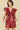 A woman modeling a sleeveless, deep burgundy faux-leather mini dress with dramatic ruffled shoulder details and a gathered front, set against a solid beige background.