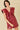A woman modeling a sleeveless, deep V-neck mini dress in a glossy burgundy faux leather material, featuring dramatic ruffled shoulder details and a gathered waist, set against a solid beige background.