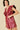 A woman modeling a shiny burgundy faux-leather mini dress with dramatic ruffle details on the shoulders and a gathered waist, set against a solid beige background.