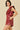 A woman modeling a sleeveless burgundy faux-leather mini dress with a large ruffled bow detail on the chest and side cutouts, posed against a solid beige background.