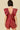 A woman viewed from the back wearing a shiny red sleeveless romper with dramatic ruffled shoulder details and a deep open back, against a plain beige studio background.