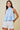 A woman modeling a light blue sleeveless halter top with a ruffled hem, paired with white shorts, against a solid beige background.