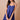 A woman modeling a royal blue sleeveless mini dress with spaghetti straps, a gathered sweetheart neckline, and a button-front bodice, posed against a solid beige studio background while holding a tan handbag.
