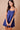 A woman modeling a royal blue sleeveless mini dress with spaghetti straps, a gathered sweetheart neckline, and a button-front bodice, posed against a solid beige studio background while holding a tan handbag.