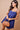 A woman modeling a royal blue sleeveless mini dress with thin straps, a gathered sweetheart neckline, and a flared skirt, holding a tan structured handbag against a solid beige studio background.