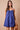 A woman modeling a sleeveless blue mini dress with thin straps, a gathered sweetheart neckline, and a button-front bodice, standing against a plain beige studio background and holding a small brown handbag.