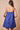 A woman shown from the back wearing a blue slip-style dress with thin straps and a loose, flared silhouette against a plain beige studio background.