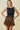 A woman modeling a sleeveless black smocked crop top with a ruffled hem and a matching smocked tiered mini skirt featuring a brown and black zebra print, set against a solid beige background.