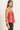 A woman wearing a shiny, metallic pink-red camisole top and black pants, photographed from the side against a plain light background.