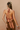 A woman shown from the back wearing a terracotta-colored open-back halter top with long tied straps and matching brown pants, standing against a textured beige wall.