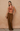 A model stands against a textured beige wall wearing a sleeveless cowl-neck top in a dusty terracotta color and matching wide-leg trousers in a medium brown. She is holding a small metallic gold clutch.