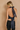 A woman shown from the back wearing a navy blue satin halter crop top with long ribbon ties and black jeans, standing against a textured beige wall.