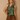 A woman modeling a sleeveless olive green satin halter top with a draped cowl neckline, paired with brown suede-like drawstring shorts, set against a textured beige studio backdrop.