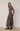 A woman stands in profile wearing a ribbed brown mock-neck long-sleeve top and a high-waisted, pleated maxi skirt with a brown, burgundy, and gray geometric diamond pattern. She is wearing black leather ankle boots against a neutral studio backdrop.