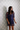 A woman wearing a sheer navy blue sequined mini dress with mesh sleeves, standing against a light gray and white patterned wallpaper background.