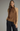 A woman modeling a sleeveless high-neck satin top in warm brown with a lace hem detail, paired with dark wide-leg pants, against a plain gray studio background.