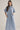A woman modeling a light blue and white vertically striped midi shirt dress with short sleeves, a collar, and side pockets, set against a plain beige studio background.