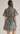 A woman shown from the back wearing a short-sleeve high-neck dress with a teal base and an allover floral print in red, orange, and cream. The dress has a matching waist tie and is photographed against a plain light gray studio background.