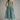 A woman modeling a sleeveless midi dress with thin straps and a flared skirt, featuring a geometric pattern of turquoise and dark brown squares, against a neutral gray studio background.