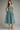 A woman modeling a sleeveless midi dress with thin straps and a flared skirt, featuring a geometric pattern of turquoise and dark brown squares, against a neutral gray studio background.