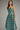 A woman modeling a sleeveless midi dress with thin straps and a blue-green and dark brown checkerboard pattern, photographed against a plain gray studio background.