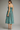 A woman modeling a sleeveless midi dress from the back, featuring a turquoise and dark brown geometric grid pattern, against a neutral gray studio background.