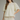 A woman modeling a cream-colored eyelet blouse with a high ruffled collar, short puff sleeves, and a gathered peplum-style hem, paired with cream trousers against a neutral gray studio background.