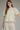 A woman modeling a cream-colored eyelet blouse with a high ruffled collar, short puff sleeves, and a gathered peplum-style hem, paired with cream trousers against a neutral gray studio background.