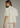 A woman shown from the back wearing a cream-colored eyelet blouse with puff sleeves and a high mock neck, paired with light beige trousers, against a solid gray studio background.