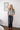 A woman stands indoors against a white shiplap wall wearing a gray short-sleeve knit top with white polka dots and dark wide-leg jeans. Two framed fish prints hang on the wall behind her.