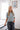 A woman with blonde hair stands in a bedroom wearing a gray short-sleeve knit sweater with white polka dots and dark gray jeans.