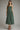 A woman modeling a sage green pleated midi dress with spaghetti straps and a gathered waist, paired with black heeled sandals against a neutral gray studio background.
