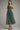 A woman modeling a sage green pleated midi dress with spaghetti straps and side pockets, paired with black heeled sandals, against a neutral gray studio background.