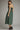 A woman modeling a sage green midi dress with thin spaghetti straps, a smocked bodice, and a gathered skirt, shown from the back against a neutral gray studio background.