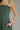 A close-up of a woman wearing a dark green pleated spaghetti-strap dress, showing the side profile, hand in pocket, and a beige textured cuff bracelet against a solid gray background.