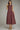 A woman modeling a sleeveless, knee-length A-line dress in a deep burgundy color with a white polka dot pattern, paired with gold flat shoes, against a neutral gray studio background.