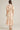 A woman viewed from the back wearing a light blush pink midi dress with a soft floral print, short puff sleeves, and a matching waist tie, paired with beige heels against a neutral studio background.