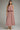 A woman modeling a sleeveless midi dress with a gathered waist and an allover small red geometric pattern on a dusty pink background, paired with flat tan shoes, against a solid gray studio background.