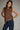 Woman wearing a brown sleeveless top with decorative trim and blue jeans against a gray background