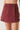 A close-up of a model wearing a high-waisted, wrap-style mini skirt in a washed burgundy denim fabric, shown from the waist down against a plain light background.