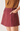 A close-up of a model wearing a dusty rose denim mini skirt with a button closure and side pockets, paired with a brown top against a plain light background.