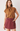 A woman modeling a sleeveless mustard-brown top with a large front bow tie and a dusty rose denim mini skirt, photographed against a clean off-white studio background.