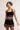 A woman modeling a matching black and white athletic set consisting of a ribbed sports bra, a high-waisted tennis skirt with white stripes, and a black headband with a small white logo, against a plain light gray studio background.