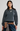 A woman modeling a dark gray crewneck sweatshirt with unique horizontal seam details on the sleeves, paired with blue jeans and a brown plaid shirt tied around her waist, against a light gray studio background.