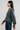 A woman in profile wearing a dark gray knit sweater and blue jeans, standing against a plain light gray studio background.